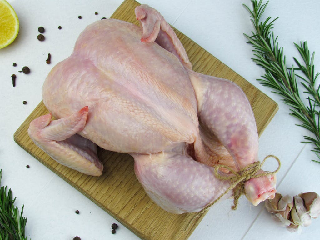 Overhead view of a raw, whole chicken with rosemary and spices on a wooden board.